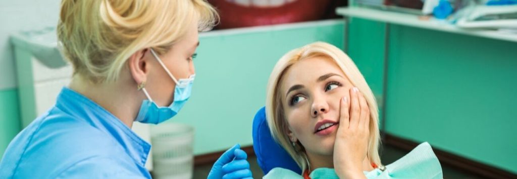 A dental hygienist in a mask talking to a concerned female patient in the treatment chair about her dental emergency
